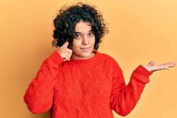 Fototapeta na wymiar Young hispanic woman with curly hair wearing casual winter sweater confused and annoyed with open palm showing copy space and pointing finger to forehead. think about it.