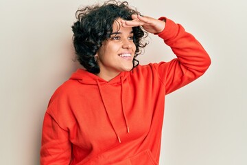 Young hispanic woman with curly hair wearing casual sweatshirt very happy and smiling looking far away with hand over head. searching concept.