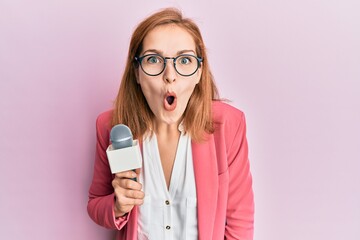 Young caucasian woman holding reporter microphone scared and amazed with open mouth for surprise, disbelief face