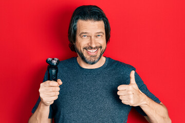 Middle age caucasian man holding electric razor machine smiling happy and positive, thumb up doing...