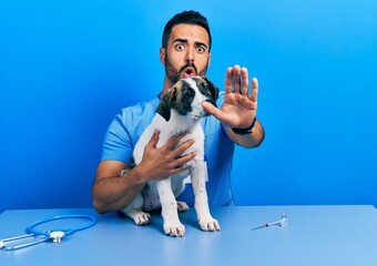 Handsome hispanic veterinary man with beard checking dog health doing stop gesture with hands...