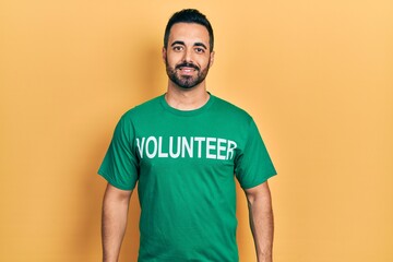 Handsome hispanic man with beard wearing volunteer t shirt looking positive and happy standing and...