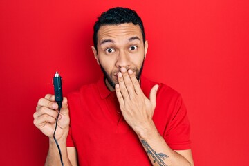 Hispanic man with beard holding car charger covering mouth with hand, shocked and afraid for...