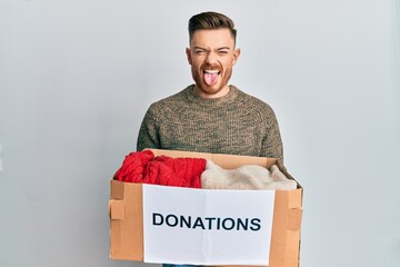 Young redhead man volunteer holding donations box sticking tongue out happy with funny expression.