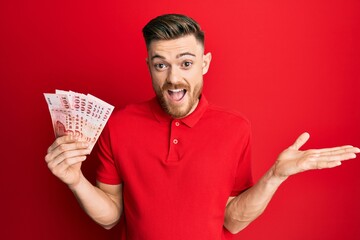 Young redhead man holding 100 new zealand dollars banknote celebrating achievement with happy smile and winner expression with raised hand