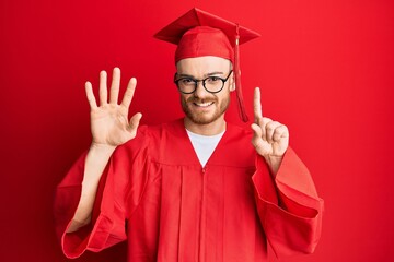 Young redhead man wearing red graduation cap and ceremony robe showing and pointing up with fingers...