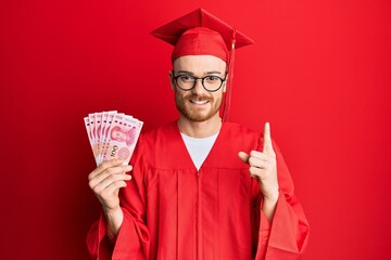 Young redhead man wearing graduation robe holding 100 yuan smiling with an idea or question...