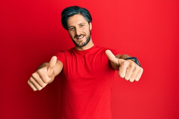 Young hispanic man wearing casual red t shirt approving doing positive gesture with hand, thumbs up smiling and happy for success. winner gesture.