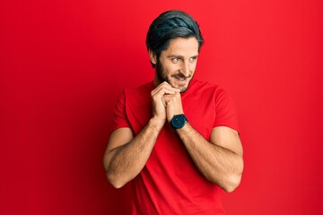 Young hispanic man wearing casual red t shirt laughing nervous and excited with hands on chin looking to the side
