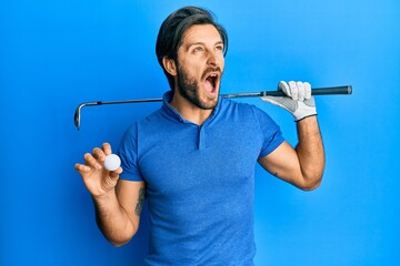 Young hispanic man holding golf ball angry and mad screaming frustrated and furious, shouting with...