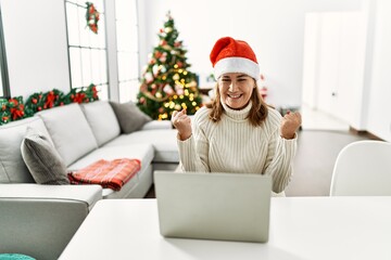 Middle age woman wearing santa claus hat using laptop screaming proud, celebrating victory and success very excited with raised arms