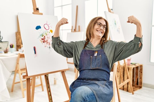 Middle age artist woman at art studio showing arms muscles smiling proud. fitness concept.