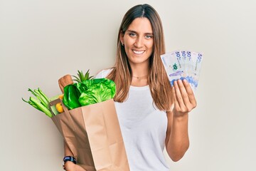 Brunette young woman holding groceries and 50 colombian pesos banknotes smiling with a happy and...