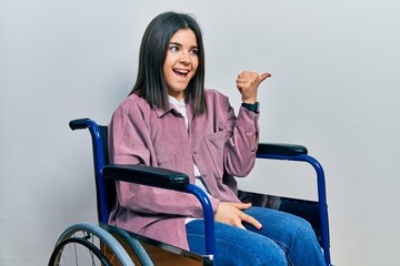 Young brunette woman sitting on wheelchair smiling with happy face looking and pointing to the side...