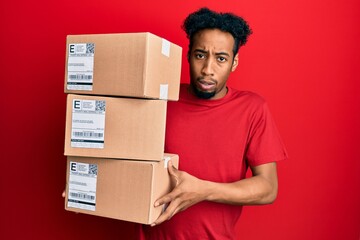 Young african american man with beard holding delivery packages in shock face, looking skeptical and sarcastic, surprised with open mouth