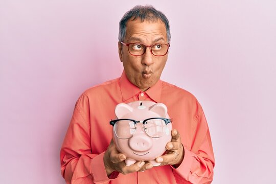 Middle age indian man holding piggy bank with glasses making fish face with mouth and squinting eyes, crazy and comical.