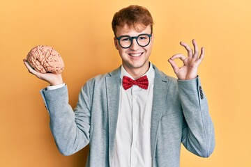 Young caucasian man wearing glasses holding brain doing ok sign with fingers, smiling friendly...