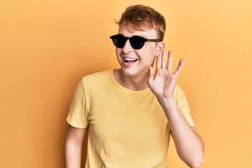 Young caucasian man wearing stylish sunglasses smiling with hand over ear listening and hearing to rumor or gossip. deafness concept.
