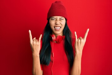 Young chinese woman listening to music using headphones shouting with crazy expression doing rock...