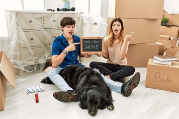 Young caucasian couple with dog holding our first home blackboard at new house surprised pointing with finger to the side, open mouth amazed expression.