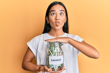 Young asian woman holding jar with savings making fish face with mouth and squinting eyes, crazy...