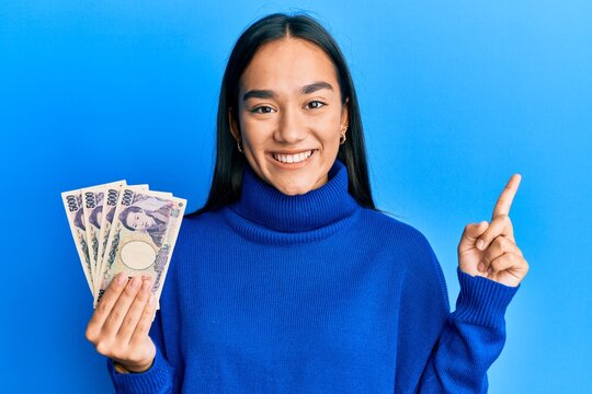 Young asian woman holding 5000 japanese yen banknotes smiling happy pointing with hand and finger to the side