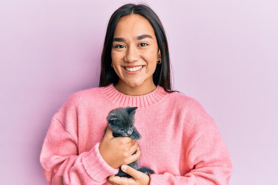 Young hispanic girl smiling happy holding cute cat over isolated pink background.