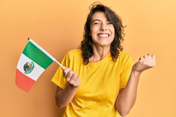 Young hispanic woman holding mexico flag screaming proud, celebrating victory and success very excited with raised arm