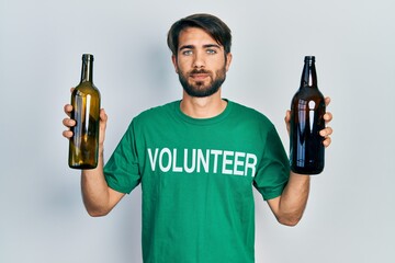 Young hispanic man wearing volunteer t shirt holding recycling bottle glass relaxed with serious...