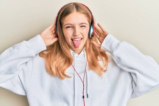 Beautiful young caucasian girl listening to music using headphones sticking tongue out happy with funny expression.