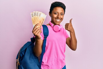 Young african american girl wearing student backpack holding norwegian krone banknotes smiling happy and positive, thumb up doing excellent and approval sign