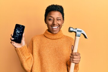 Young african american girl holding broken smartphone showing cracked screen and hammer winking looking at the camera with sexy expression, cheerful and happy face.