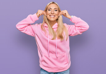 Young blonde woman wearing casual sweatshirt smiling pulling ears with fingers, funny gesture....