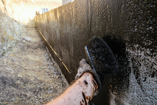 Protective waterproofing coating of walls in contact with the ground with bituminous mastic. Worker hand  applies bitumen mastic on the foundation.