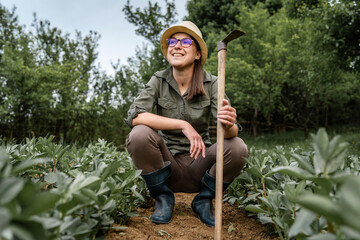 Front view of young caucasian happy woman female farmer in the agriculture filed holding hoe while having a rest taking a brake from work - real people horticulture and self sufficiency concept