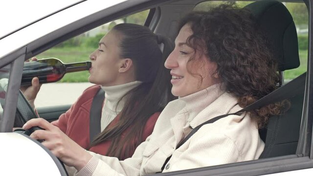 Two girlfriends drink wine from a bottle while sitting in the car. Drunk driving.