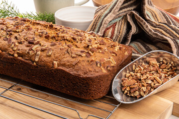 A loaf of banana bread with pecan pieces baked in fresh from the oven on a wire cooling rack with a baker's scoop of pecan pieces
