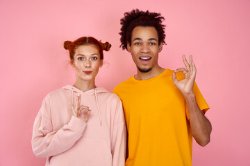 Mixed race couple of European girl and African American guy showig ok sign with fingers, their looks expressing excitement, amazement, joy and happiness on pink studio background. Good choice of place