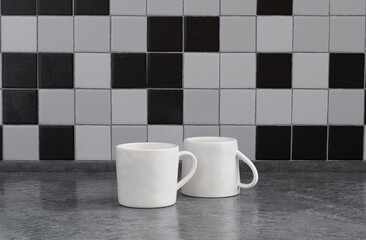 Two white porcelain coffee cups side by side on black amd white color front view kitchen counter top with faience mosaic tessellated wall, 3d Rendering, close-up view