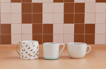 Three different coffee mugs side by side on kitchen countertop with wall, close up front view, 3d rendering, no people