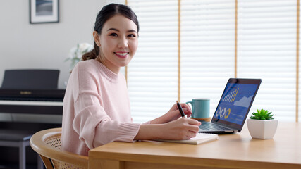 Portrait headshot young attractive beautiful asia female sit on desk smile to camera work at home remotely computer laptop job. College student, employee feel cozy happy in weekend work life balance.