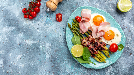 Fototapeta na wymiar Breakfast fried eggs, asparagus wrapped in bacon, ham, tomatoes and salad. Healthy balanced food. Top view, above, banner