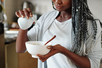 Cropped image of young woman pouring fresh milk in bowl with cornflakes or granola