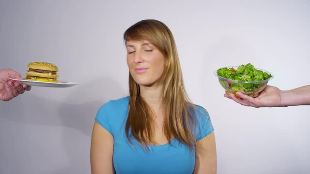 PORTRAIT, CLOSE UP: Smiling Caucasian female holds up a salad bowl after deciding what to eat for dinner. Thoughtful young woman chooses a healthy salad over a burger while weighing her lunch options.