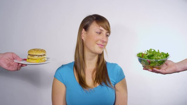 PORTRAIT, CLOSE UP: Beautiful Caucasian female opts to eat a healthy green salad over a fatty double hamburger. Thoughtful young woman chooses a salad over a burger while weighing her lunch options.