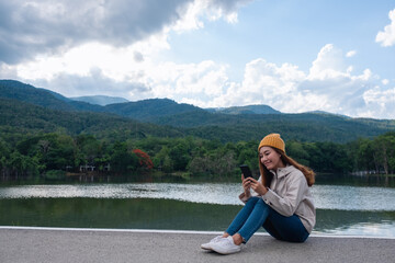 A young asian woman holding and using mobile phone while sitting by the lake and mountains