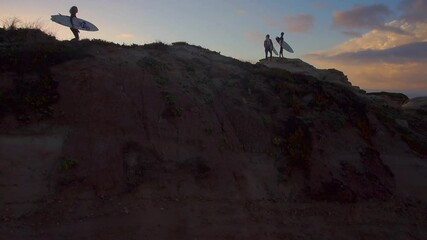 AERIAL, LENS FLARE: Fit young tourists carry their surfboards to a viewpoint on the shoreline of Portugal on a sunny summer evening. Flying over group of surfers walking along a sandy trail at sunset.
