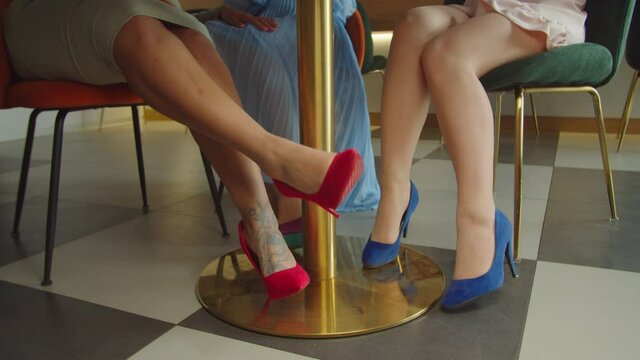 Close-up of diverse female legs in stylish high heel shoes under cafe table. Low section of woman flirting with female friend an d caressing her knee with hands during lunch in coffee shop,