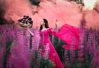 Beautiful girl in long pink dress with small ship in hand stading in flower field