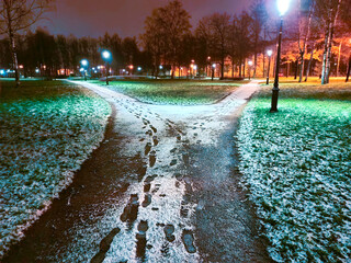 The pedestrian alley, covered with early thawed snow, branches into two directions. Night landscape in the light of street lamps.